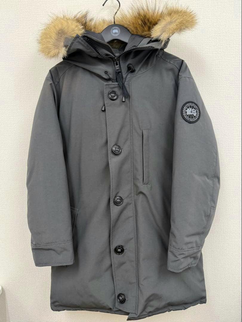 Gray CANADA GOOSE men's down jacket with a fur-trimmed hood hanging on a hanger against a wall.