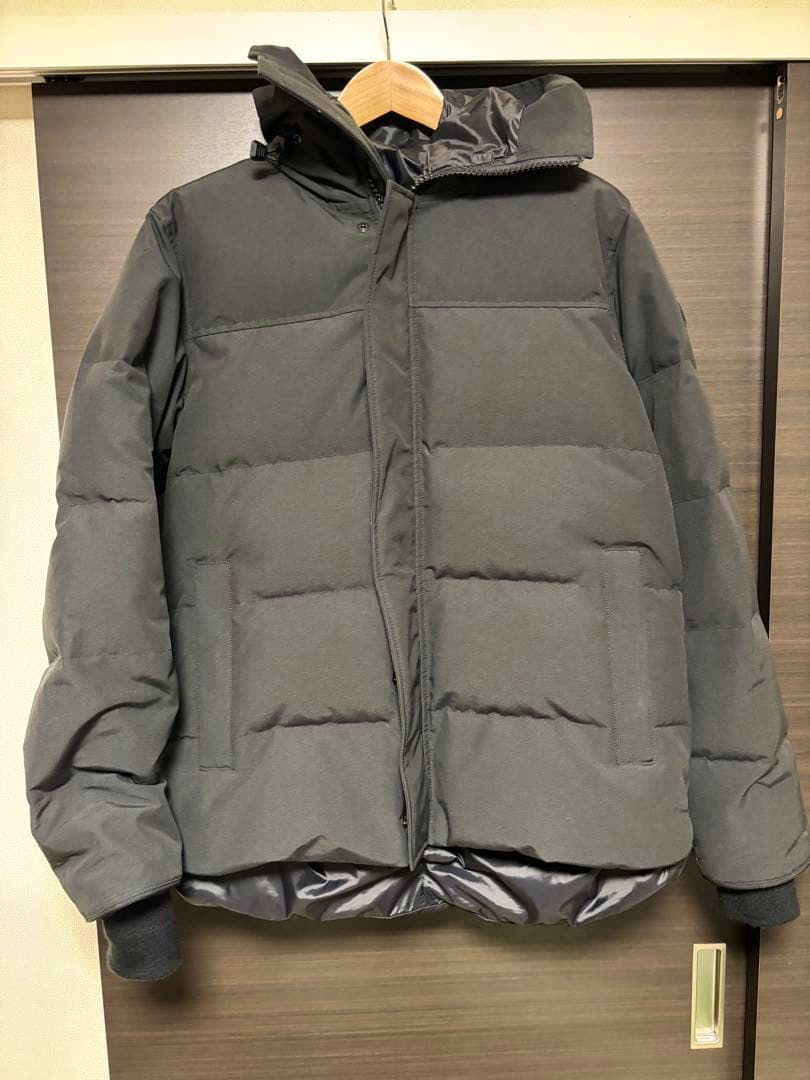 Front view of a Canada Goose men's polyester jacket hanging on a wooden hanger against a wooden door.