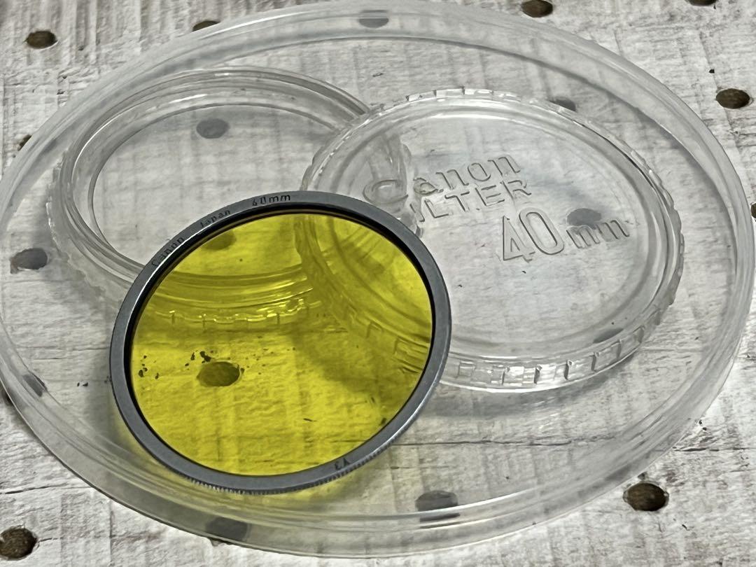 Yellow Canon 40mm lens filter with silver rim resting on a clear plastic 40mm Canon filter case.