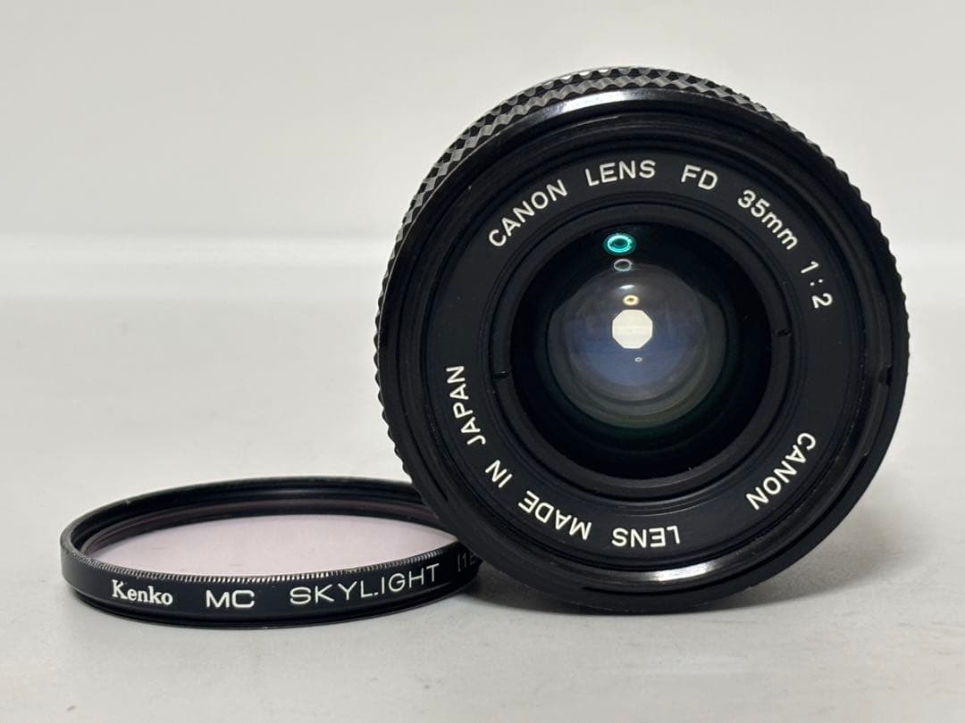 Front view of a Canon FD 35mm f/2 camera lens with a Kenko MC Skylight filter placed beside it on a light background.