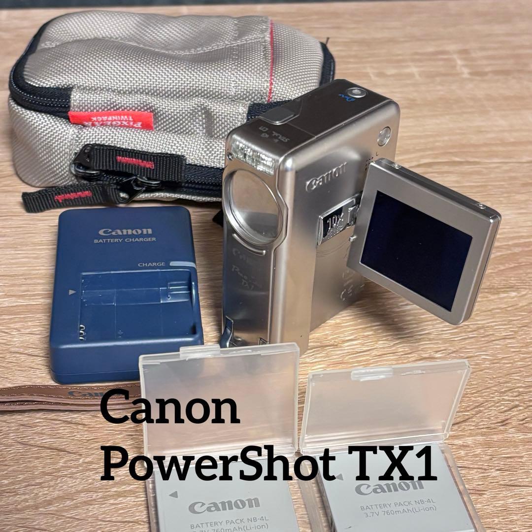 Canon PowerShot TX1 camera with its flip-out LCD, a battery charger, two battery cases, and a small carrying pouch on a wooden surface.