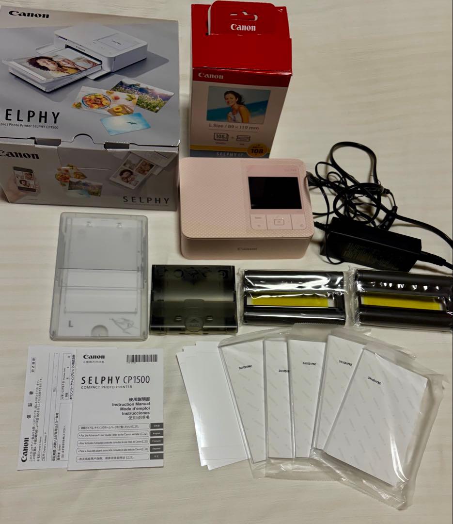 Pink Canon SELPHY CP1500 compact photo printer displayed with its box, power adapter, ink/paper packs, and manuals on a flat surface.