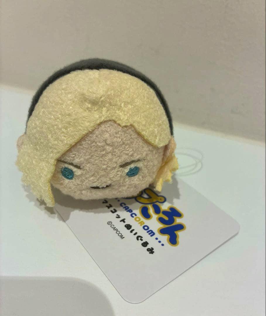 A small Capcom Capcoron バイオハザード グレース plush toy with blonde hair and blue eyes shown on a white surface with its tag.