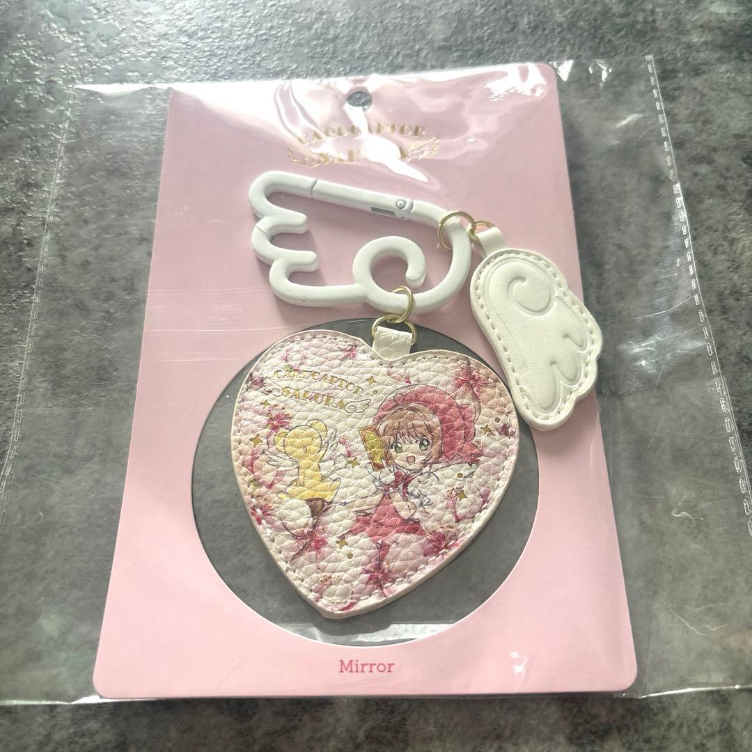 Heart-shaped compact mirror featuring Cardcaptor Sakura and Kero illustrations attached to a white wing-shaped clip, shown on pink backing in its packaging.