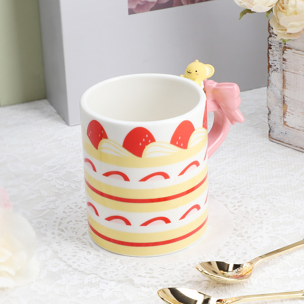 A ceramic Cardcaptor Sakura mug with a cake-pattern print and a pink bow handle topped by a small yellow Kero-chan figure.