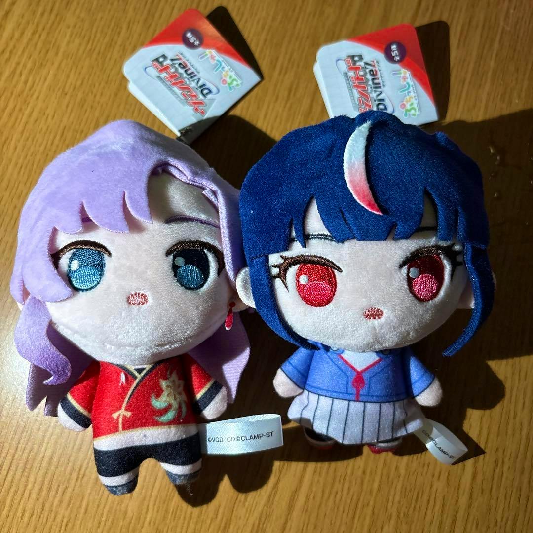 Front view of two plush mascot toys placed side by side, one with purple hair in a red outfit and one with blue hair in a blue outfit, both with attached tags.