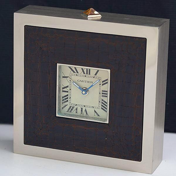 Front view of a Cartier Art Deco travel clock showing a small square white dial with Roman numerals and blue hands set in a metal case with a dark textured inset.
