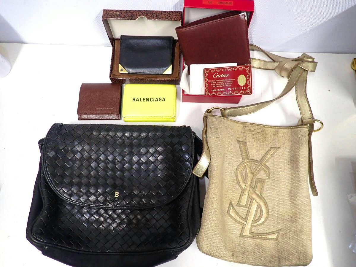 Beige Yves Saint Laurent shoulder bag with raised YSL logo shown alongside a black woven bag and several wallets on a white surface.