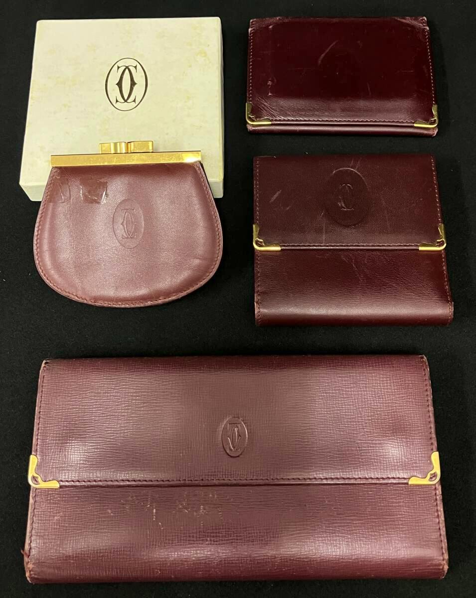 Multiple burgundy Cartier leather wallets and a matching coin purse with gold hardware arranged on a black background next to a Cartier box.