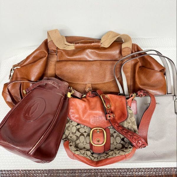 Four brand bags arranged together: a large brown tote, a Coach signature canvas bag with red-brown leather trim at center front, a burgundy leather bag at left, and a light gray tote at right.