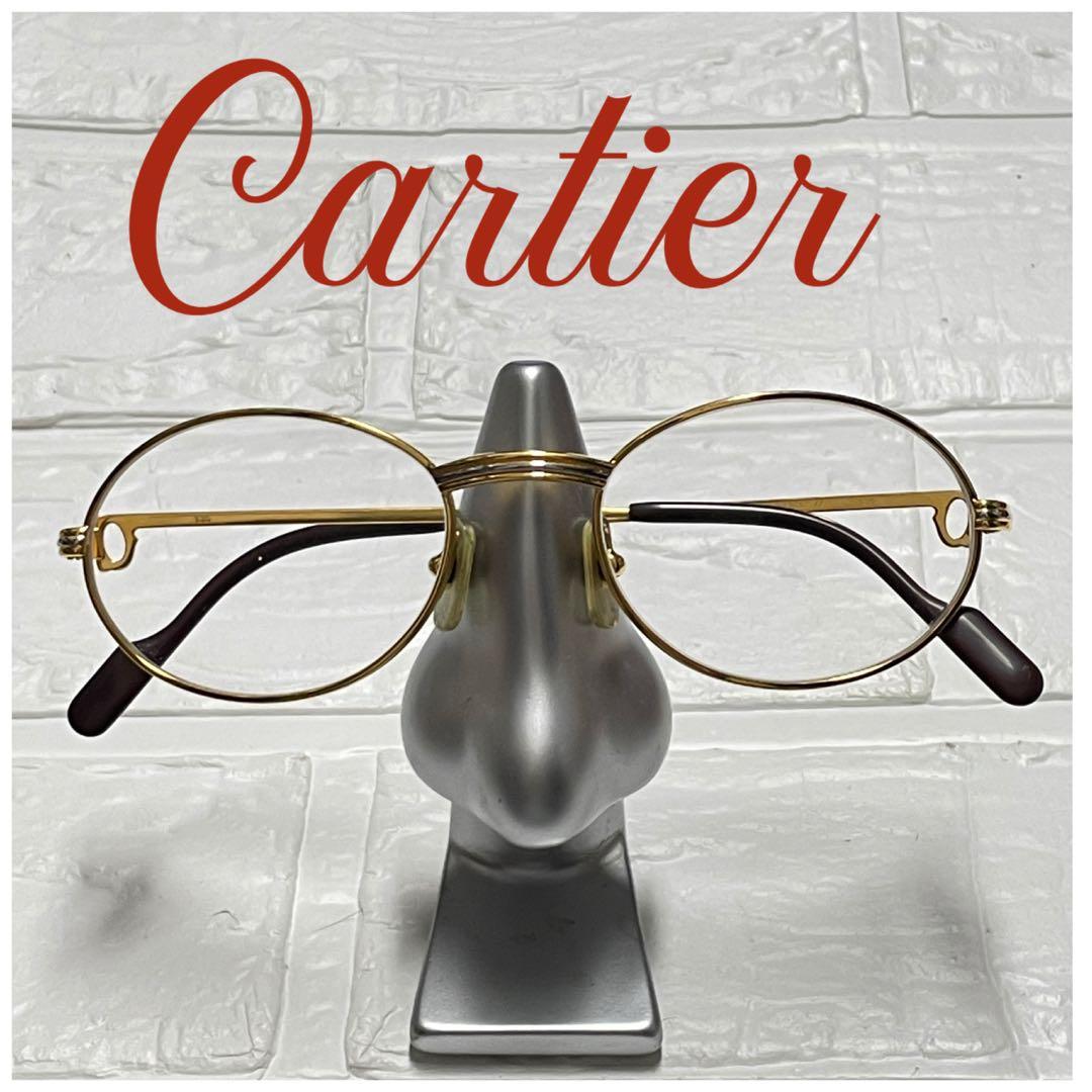 Gold-framed oval Cartier eyeglasses displayed on a silver nose-shaped stand against a white textured background.