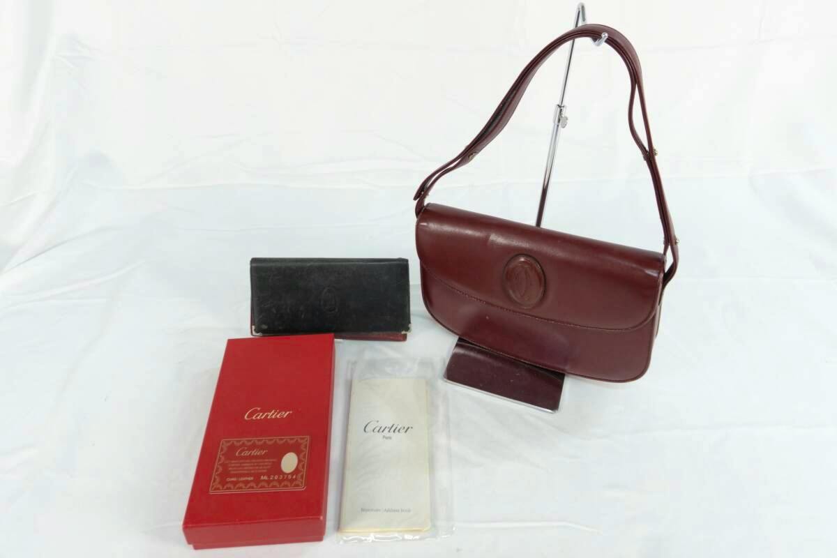 A burgundy Cartier leather shoulder bag on a display stand with a matching wallet, red Cartier box, and paperwork on a white background.