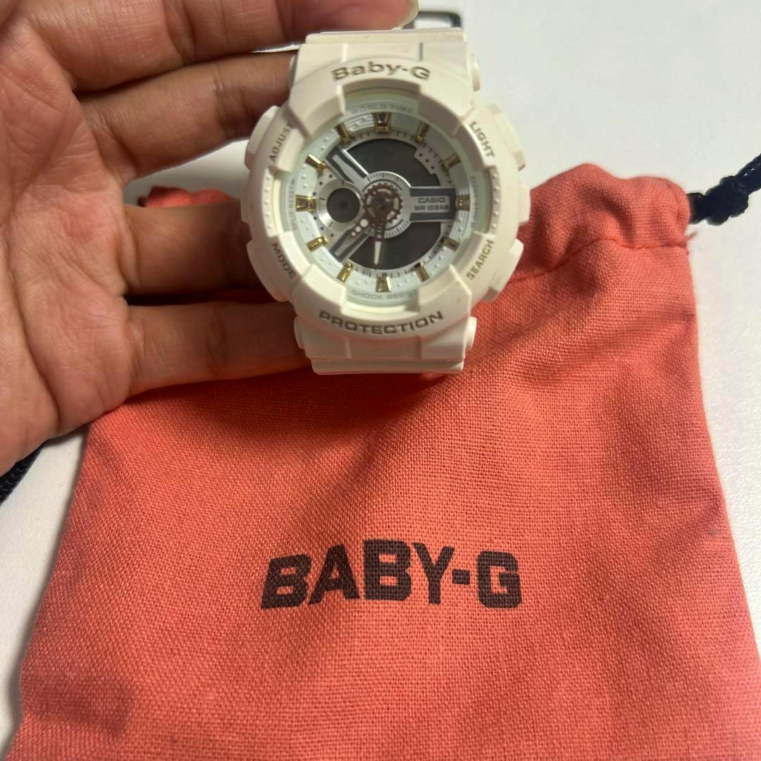 White CASIO Baby-G wristwatch held by a hand above a coral Baby-G pouch, showing the watch face.