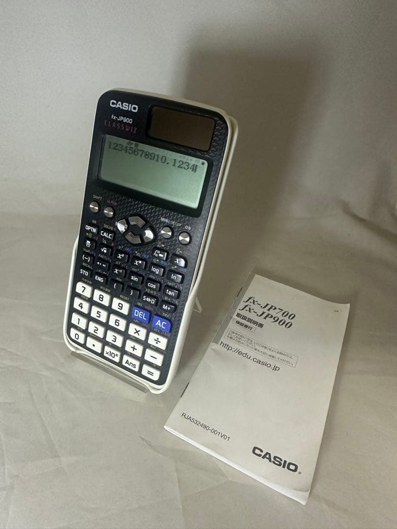 Front view of a CASIO fx-JP900 scientific calculator standing next to its instruction manual on a light surface.