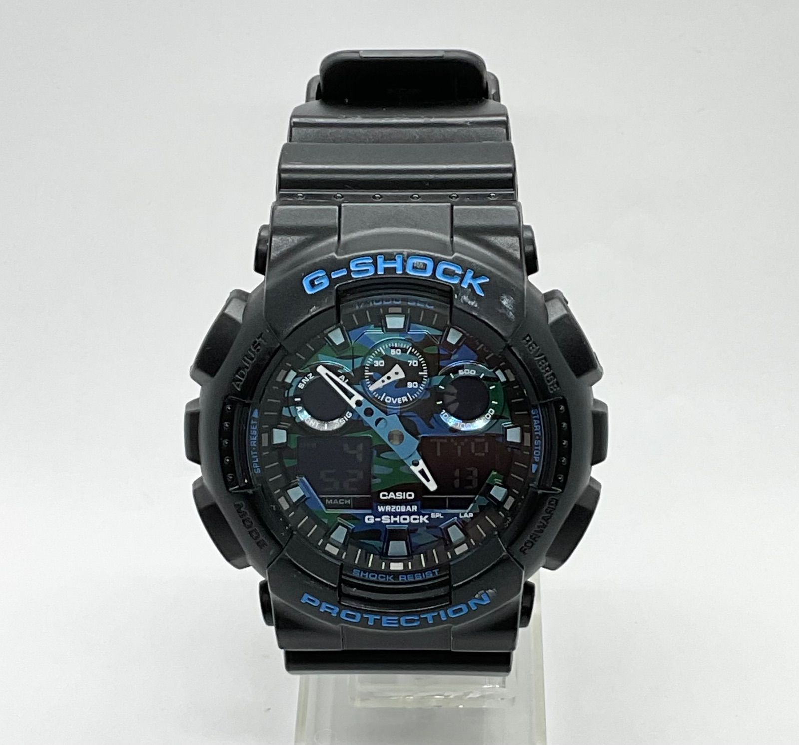 Front view of a black CASIO G-SHOCK GA-100CB camouflage analog-digital wristwatch on a clear stand.