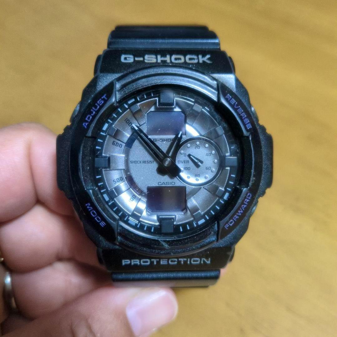Front view of a black G-SHOCK watch showing the dial, hands, and digital display held in a hand.