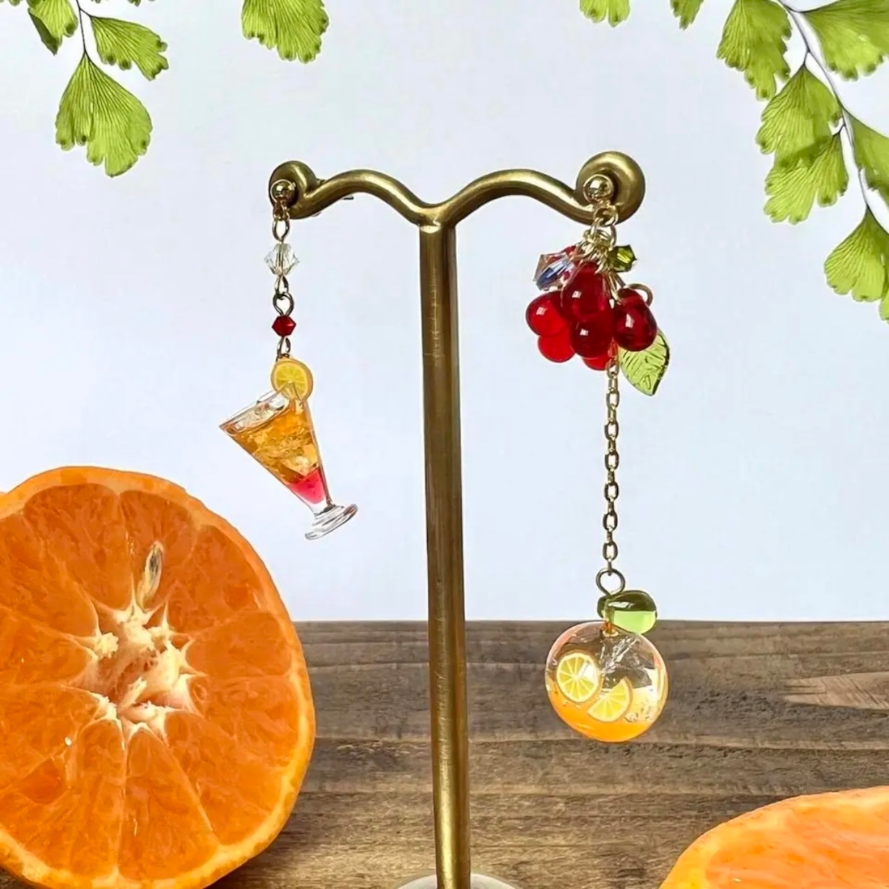 Two dangling fruit-themed earrings displayed on a metal stand with orange slices on the surface.