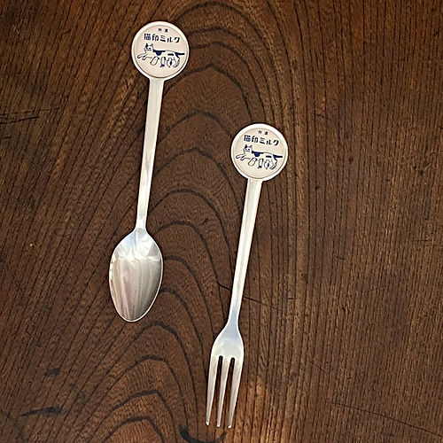 A metal spoon and fork from the "猫印ミルク" set with round tops showing cat illustrations, displayed on a wooden surface.