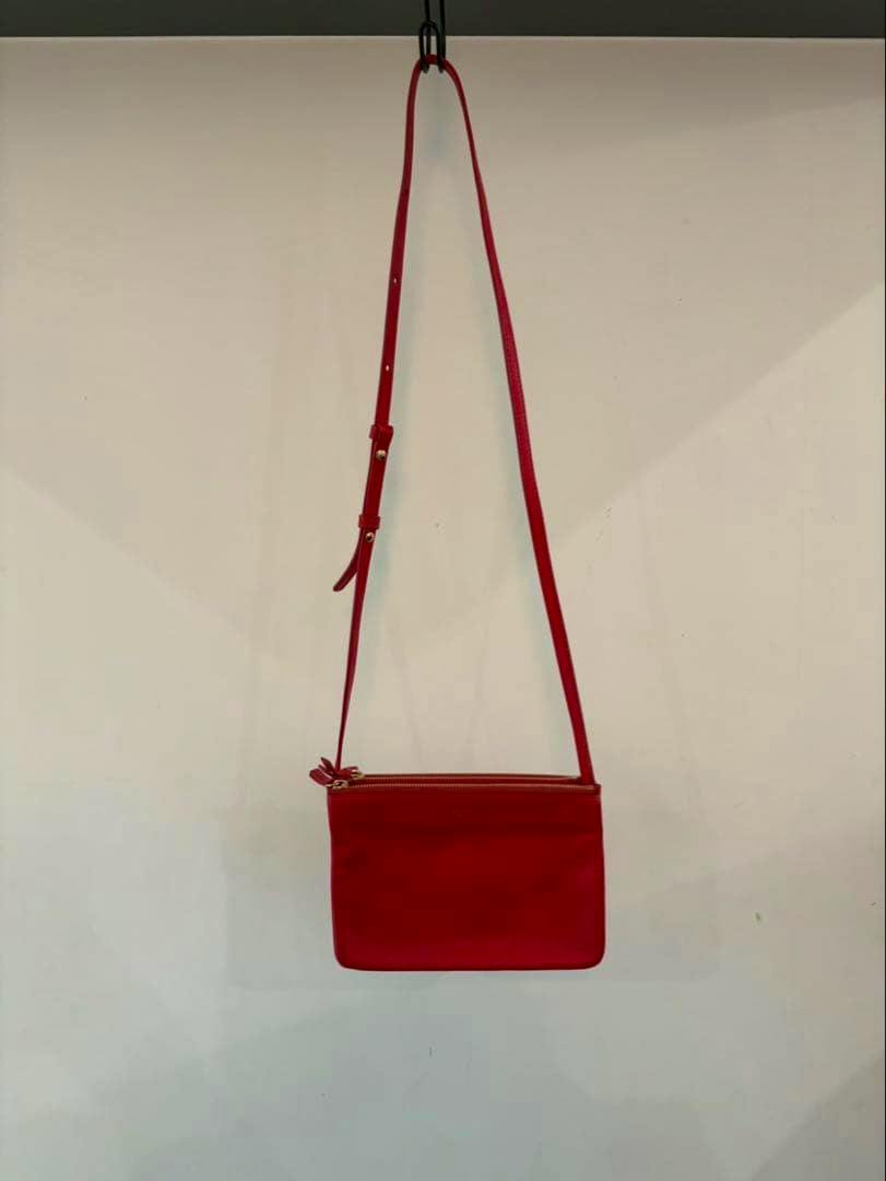 Red CELINE Trio small shoulder bag hanging from a hook against a plain wall, showing the front and long strap.