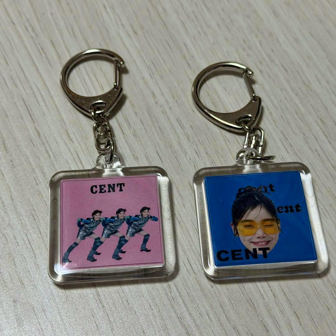 Two square acrylic keychains with metal rings on a wood surface; the left has a pink panel with three small running figures and the word “CENT,” and the right has a blue panel with a face and the word “CENT.”