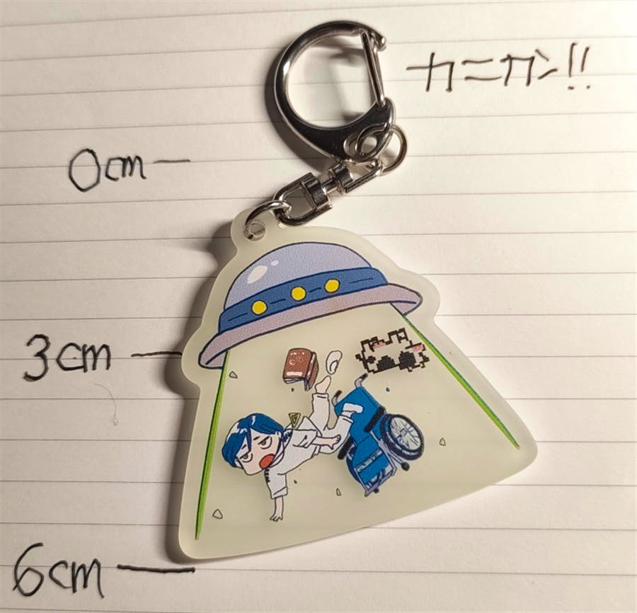 A photo of the acrylic keychain showing a UFO-shaped beam abducting a small cartoon person, placed on lined paper with 0 cm, 3 cm, and 6 cm markings.