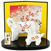 Two white ceramic horse figurines with red floral patterns displayed on a black oval base in front of a decorative folding screen and small standing plaque.