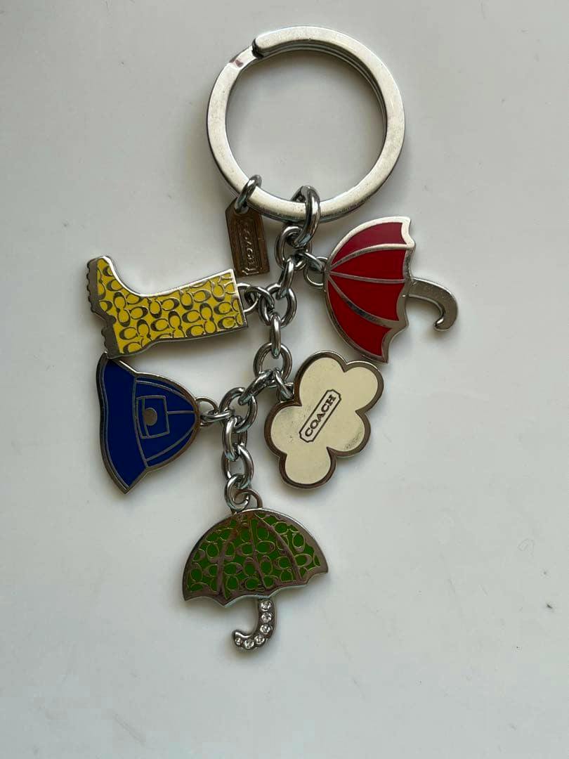 Front view of a Coach rain-themed bag charm showing a metal keyring with five colorful enamel rain-related charms (yellow boot, red umbrella, blue hat, cream flower with Coach logo, green umbrella).