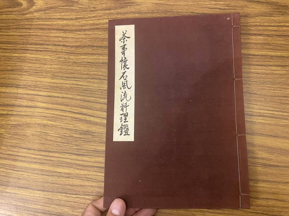 Front cover of a brown bound Japanese book with a vertical title label, held by a hand on a wooden surface.