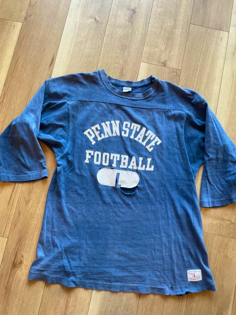 Blue men's cotton 3/4-sleeve shirt laid flat showing 'PENN STATE FOOTBALL' print on the front with a small damaged area below the graphic and a size patch at the hem.