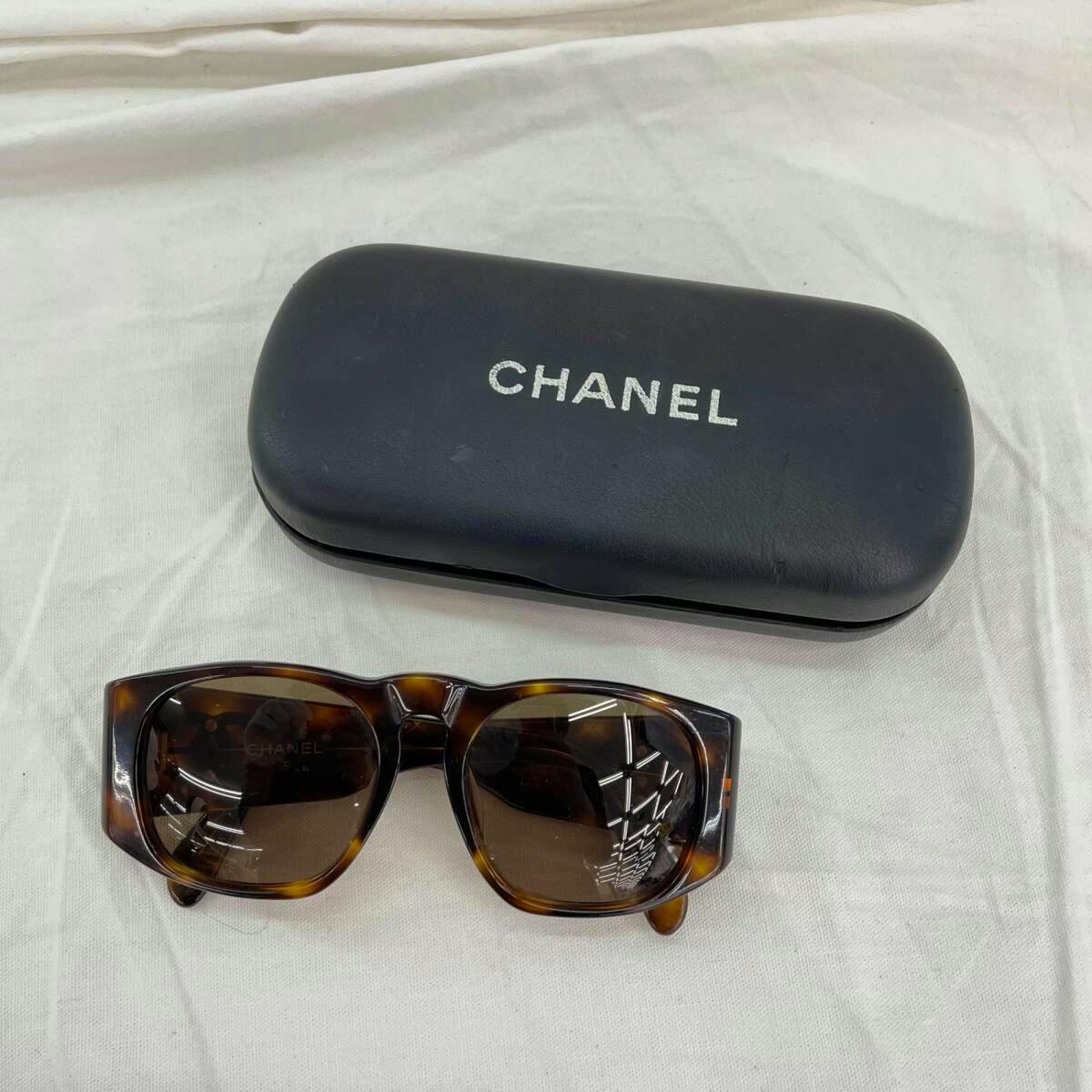 Brown tortoiseshell plastic sunglasses with brown lenses shown with a black CHANEL hard case on a white cloth background.