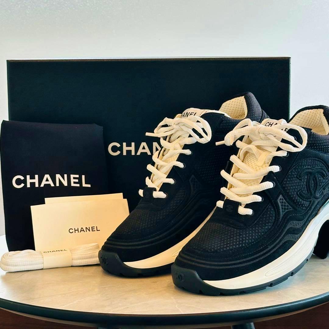 Pair of black CHANEL sneakers displayed on a table with the CHANEL box, extra laces, and documentation visible.