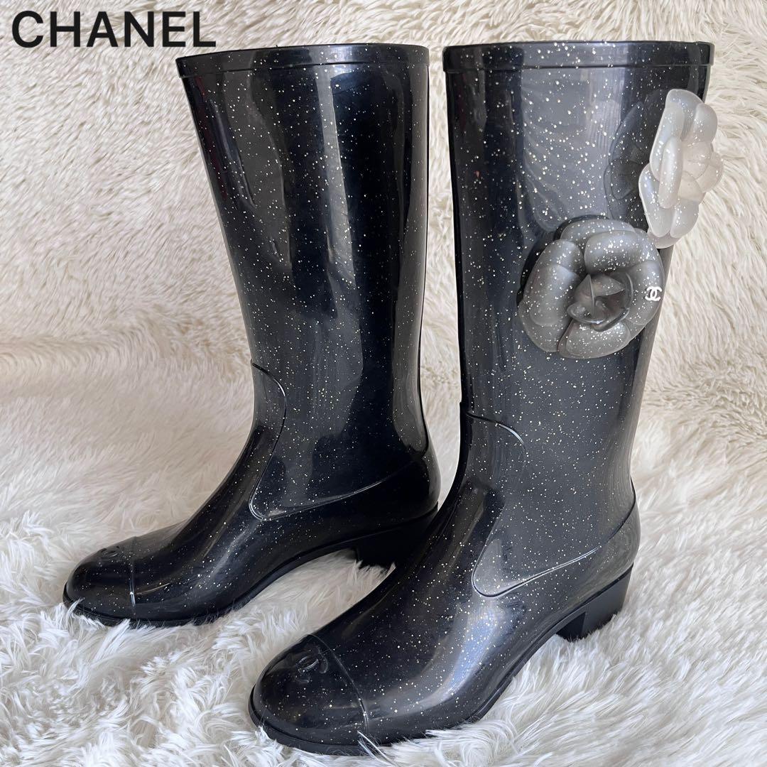 Black Chanel rain boots with silver glitter flecks and camellia flower decorations photographed on a light textured background.