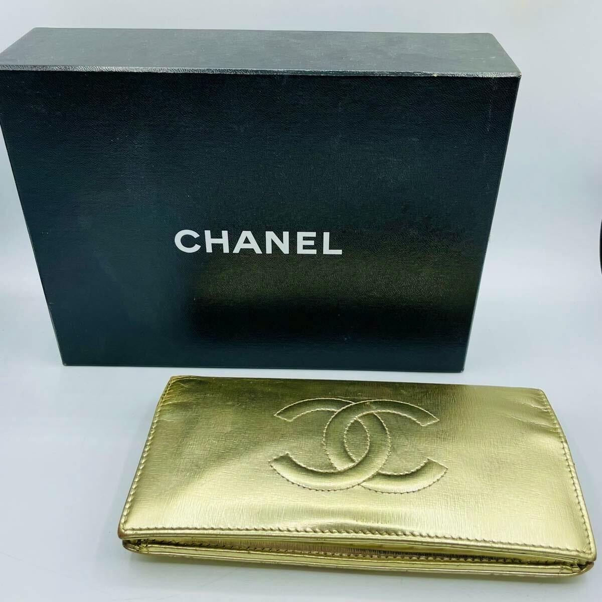 Gold CHANEL long wallet displayed in front of a black CHANEL box on a white background.