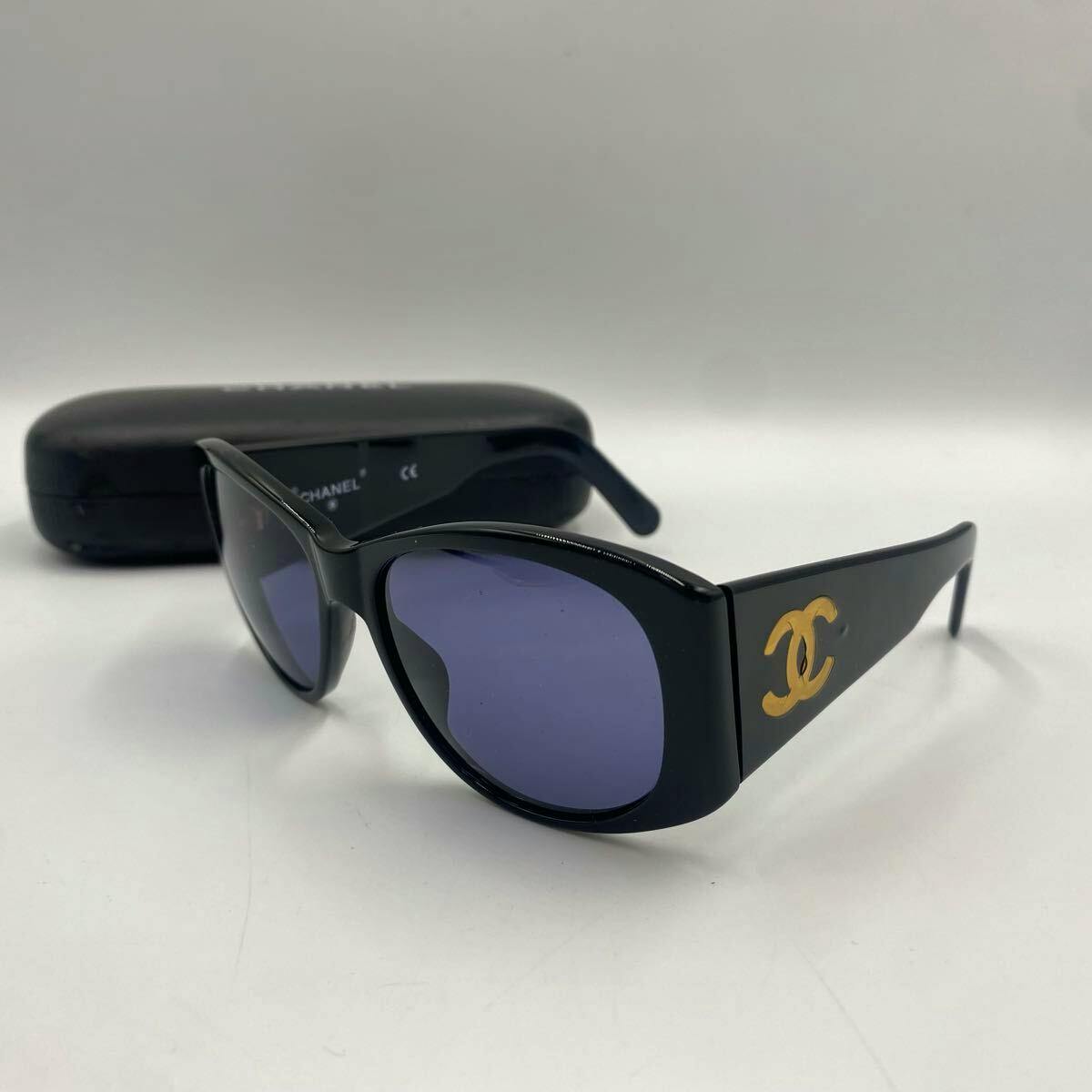 Black plastic CHANEL sunglasses with gold CC logo on the temples, with a black CHANEL case behind them.