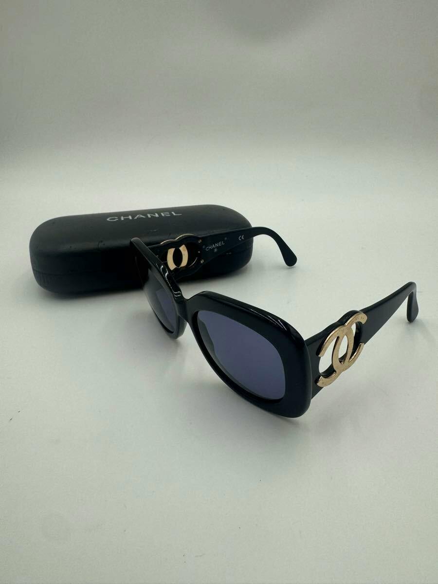 Black plastic CHANEL sunglasses with gold CC logo on the temples shown with a black CHANEL case in the background.