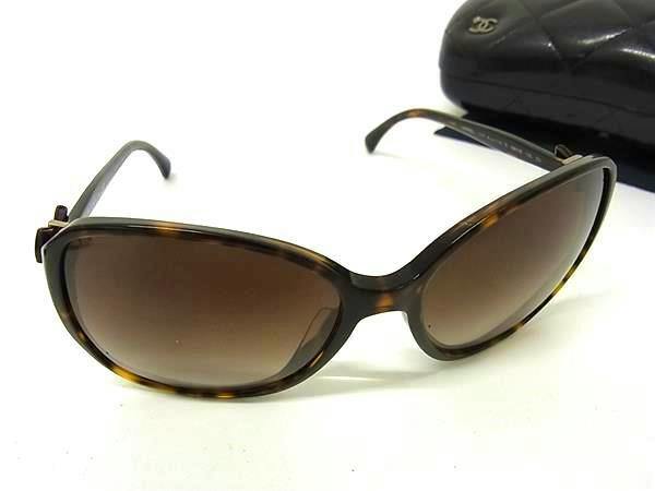 Brown tortoiseshell-patterned CHANEL women's plastic sunglasses with ribbon detail shown on a white surface with a black case in the background.