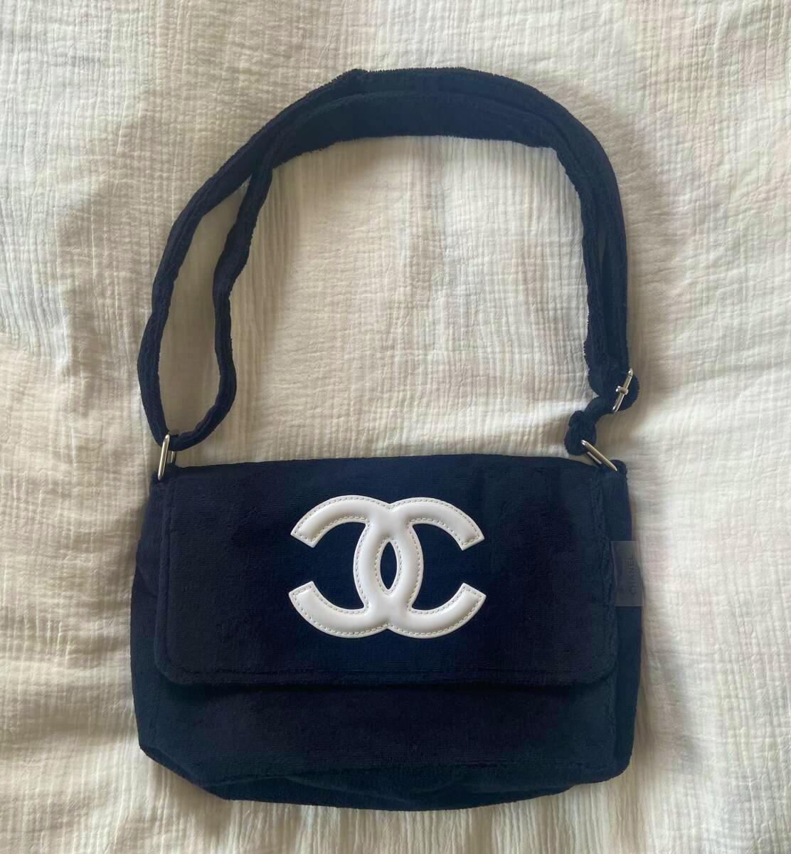 Black CHANEL shoulder bag with a white interlocking CC logo on the flap, shown flat on a white textured surface.