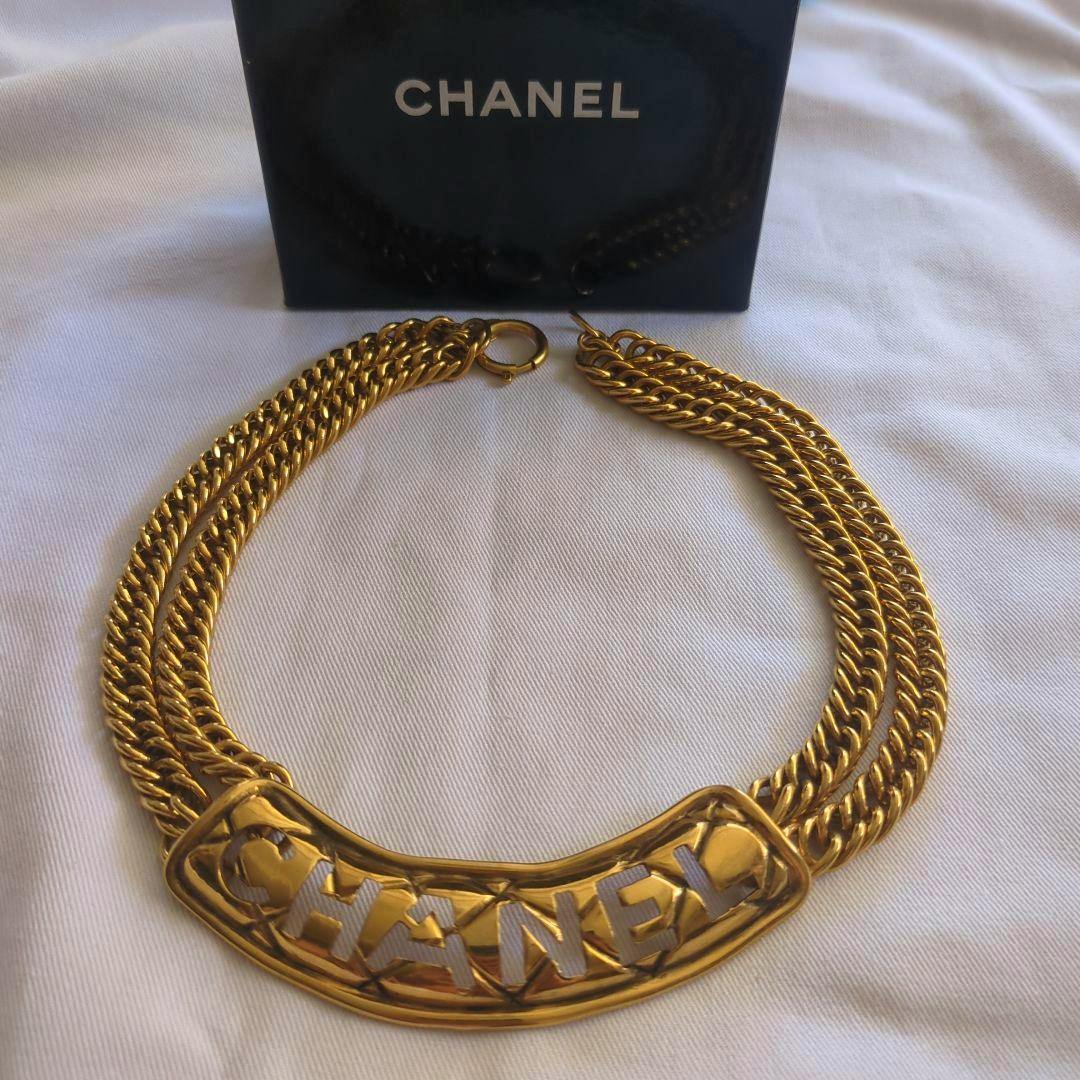 Gold-tone CHANEL necklace with a curved logo plate and triple chain laid on white fabric with a CHANEL box behind it.