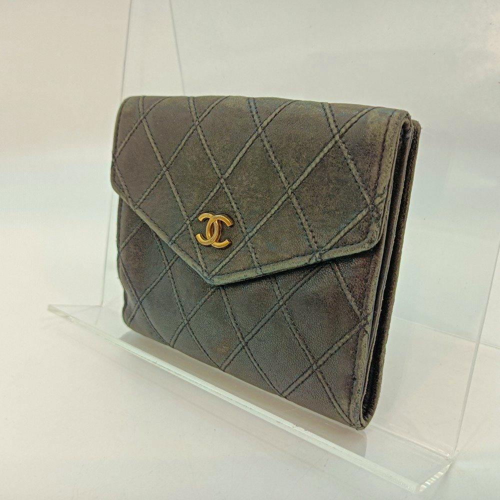Front view of the CHANEL Matelassé black bi-fold wallet with a gold CC logo on the flap, shown on a clear display stand.