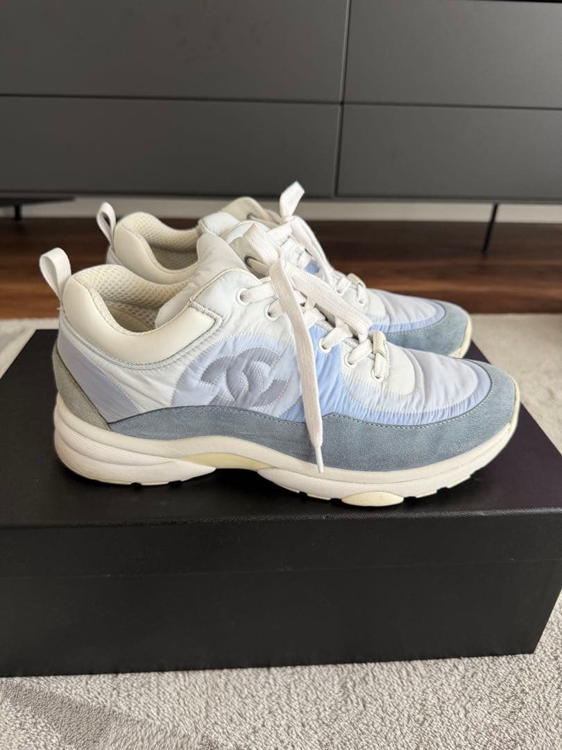 Side view of a pair of blue and white CHANEL men's nylon sneakers placed on a black box.