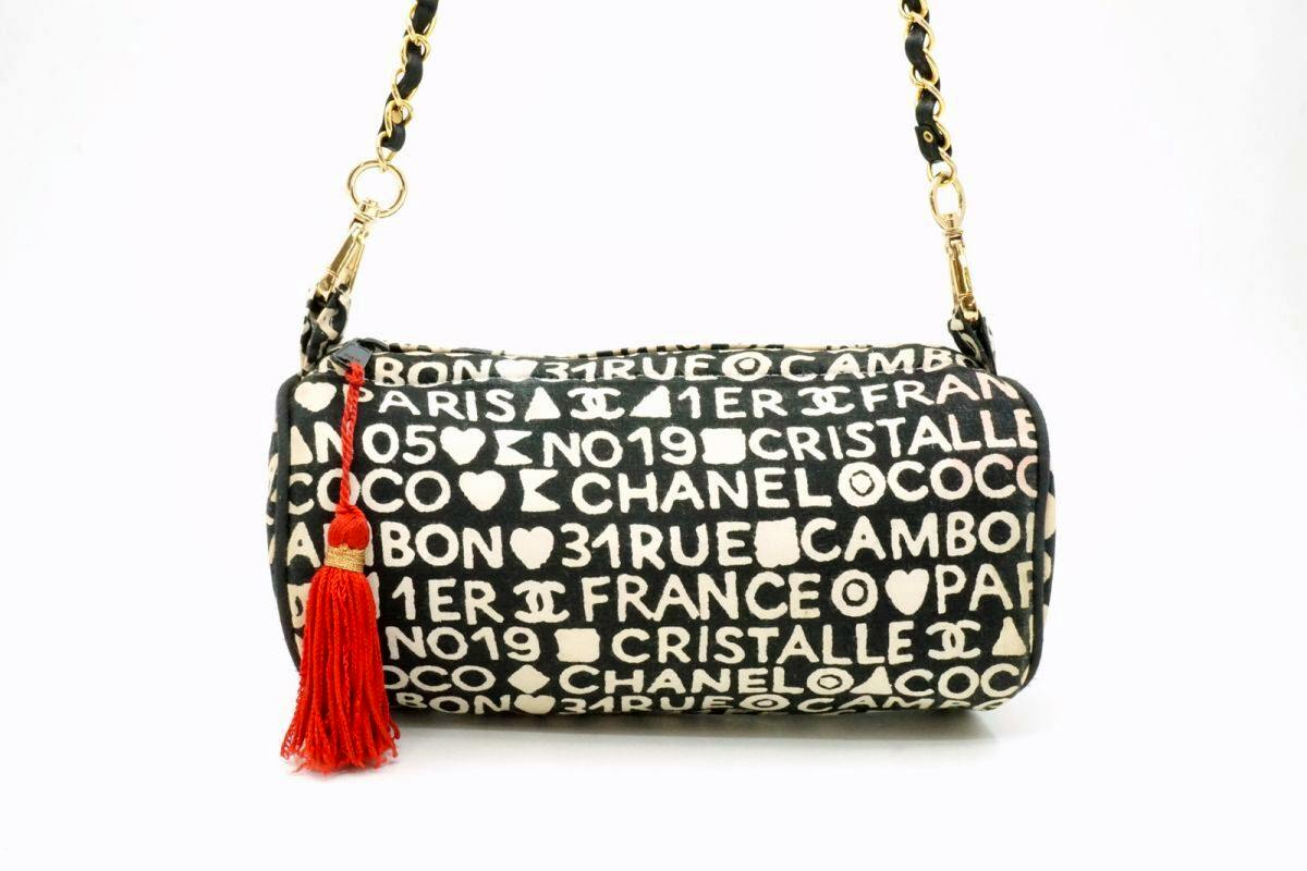 Black canvas cylindrical CHANEL mini chain shoulder bag with white logo print, gold chain strap, and a red tassel.