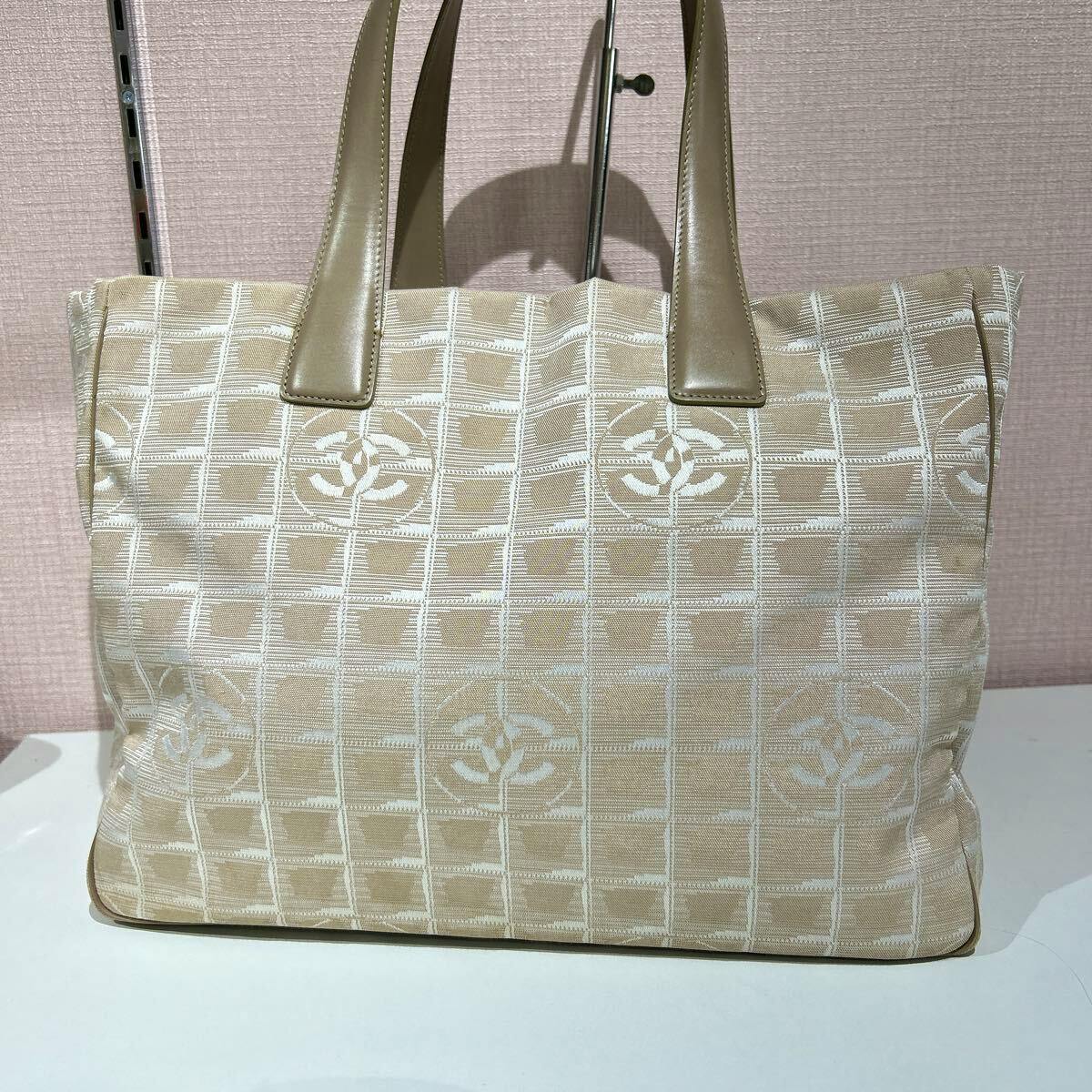Front view of a beige CHANEL New Travel Line nylon tote bag with leather handles and CC logo pattern.