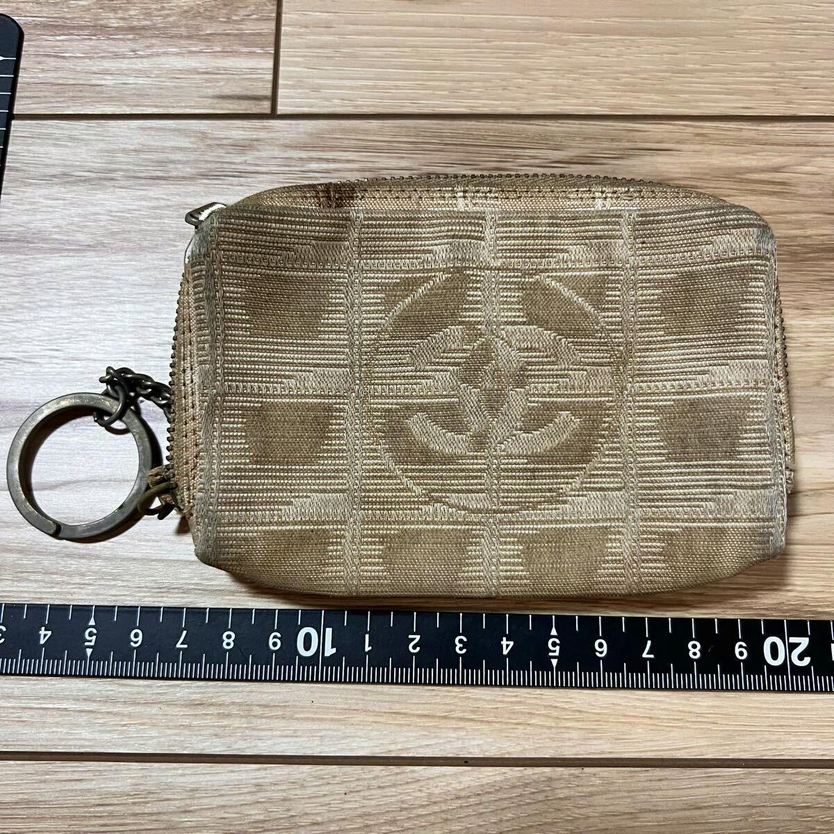 Beige CHANEL New Travel Line nylon coin case with woven CC logo and attached keyring on a wooden surface next to a measuring ruler.