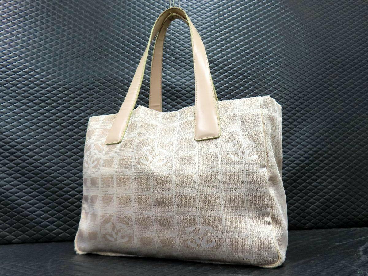 Beige CHANEL tote bag with beige leather handles and checkerboard fabric showing CC logos against a dark textured background.