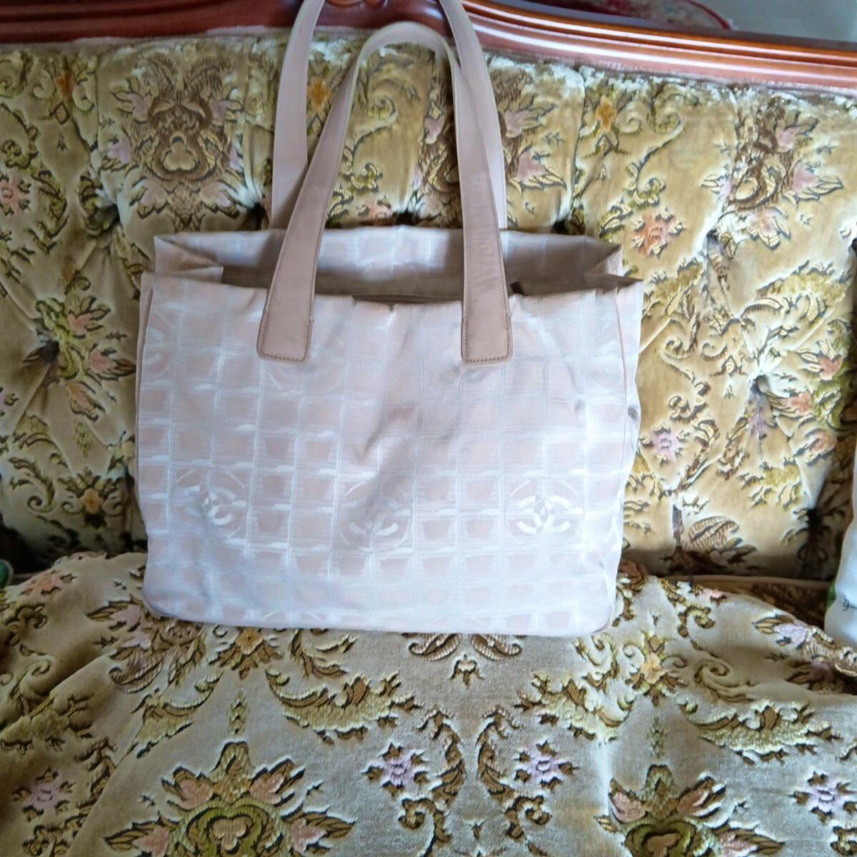 Beige CHANEL New Travel Line nylon tote bag with visible CC logo pattern shown full-front on a patterned sofa.