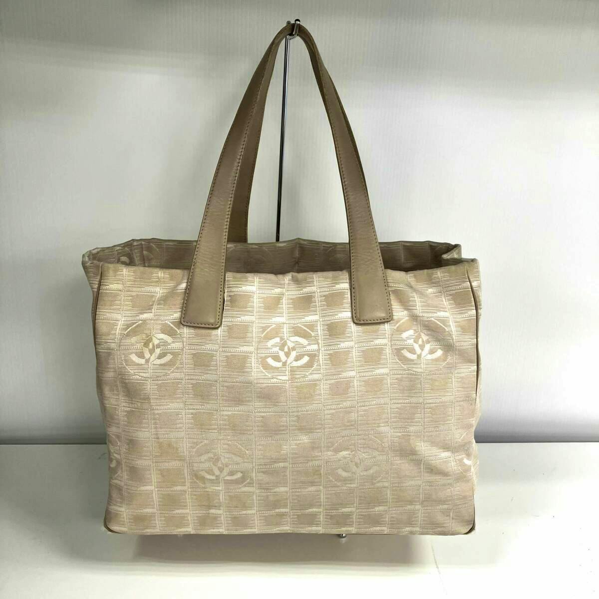 Front view of a beige CHANEL nylon New Travel Tote MM with leather handles and a repeating CC pattern, displayed on a stand.