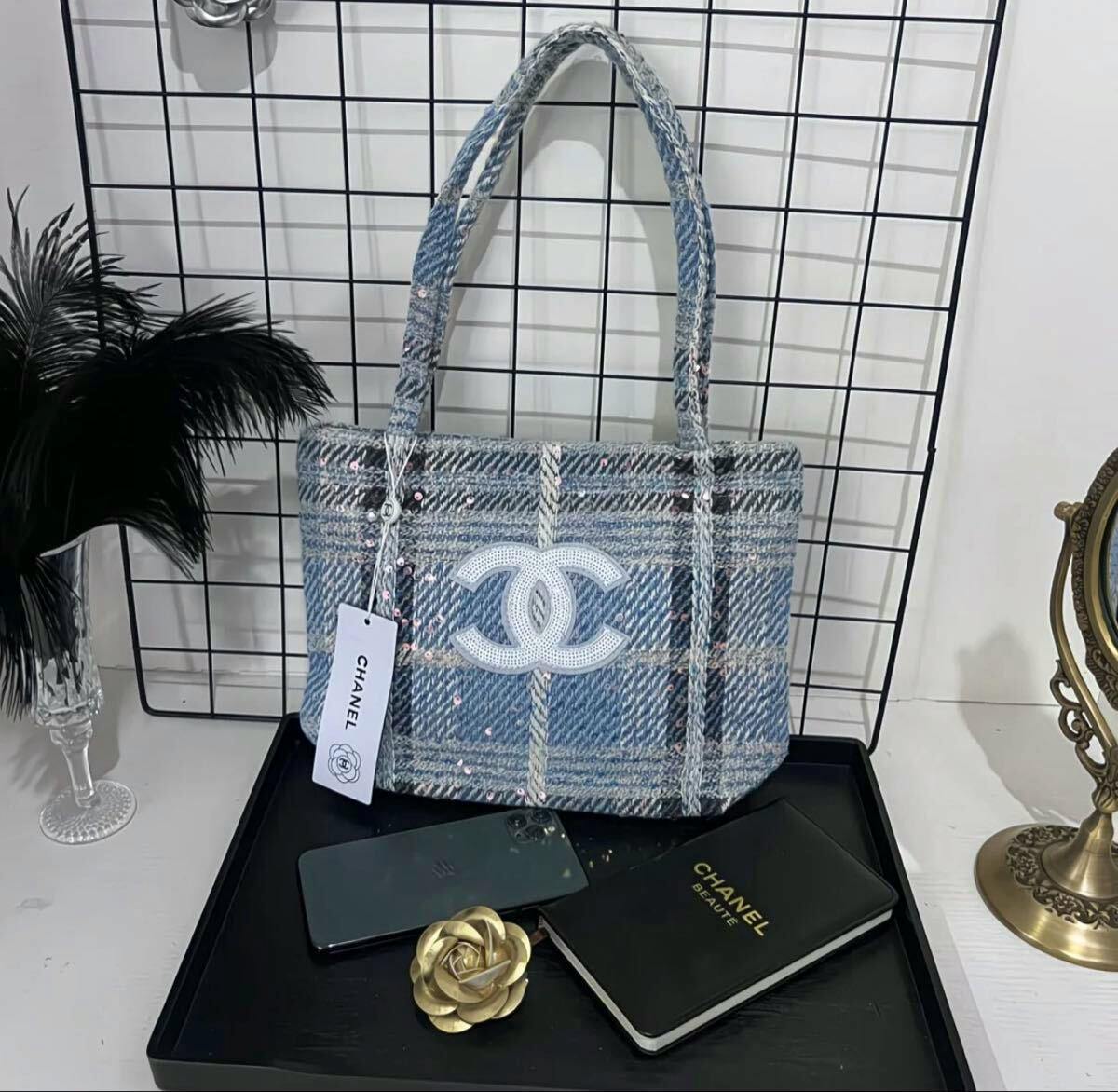 Front view of a blue patterned Chanel tote bag with an interlocking CC logo and an attached CHANEL tag displayed on a stand.