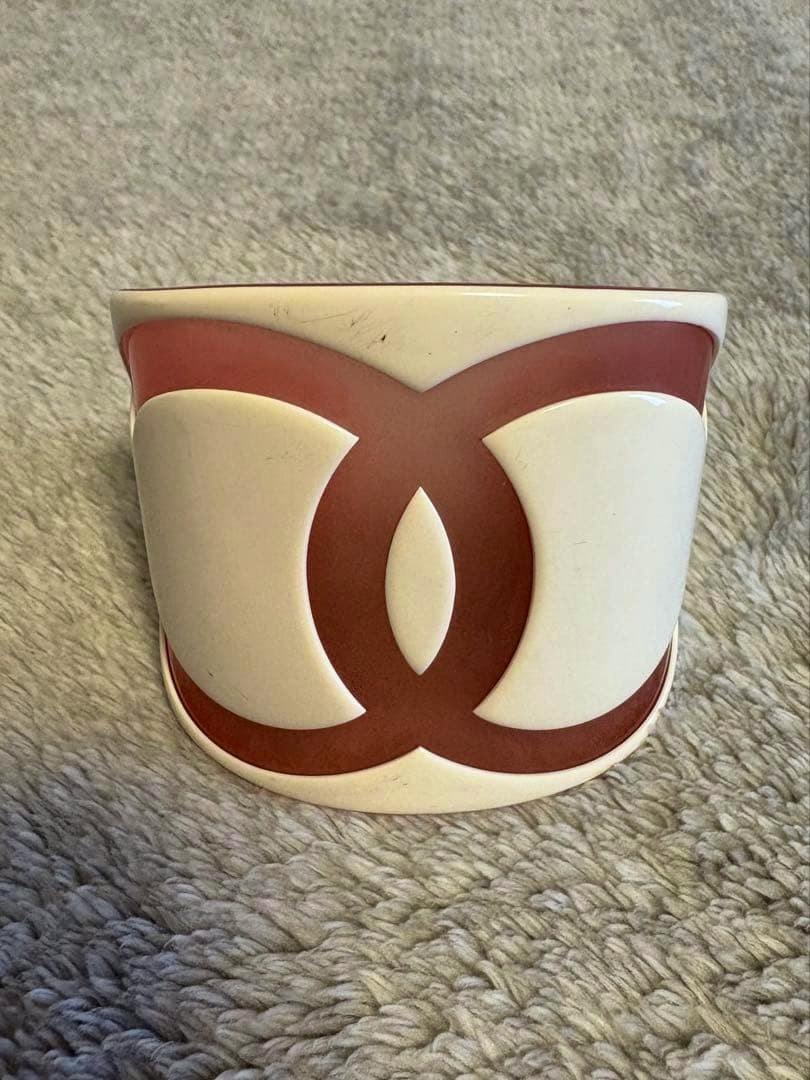 Cream-colored plastic CHANEL bangle with a large brown interlocking CC logo shown from the front on a gray textured surface.