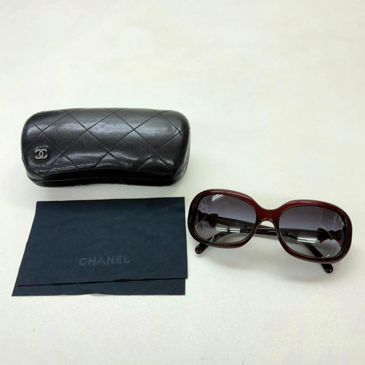 CHANEL plastic sunglasses pictured with a quilted black sunglasses case and a black cleaning cloth on a white surface.