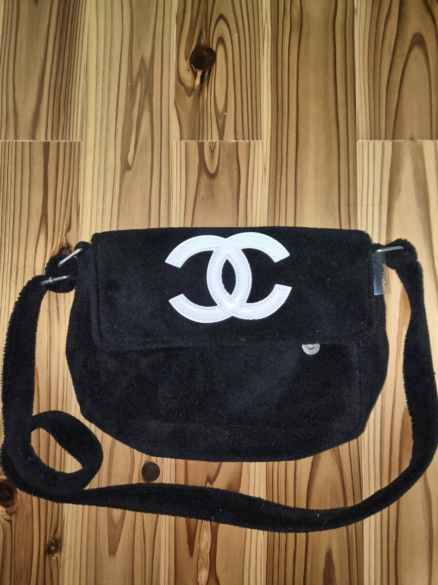 Black CHANEL pile‑fabric shoulder bag with a white interlocking CC logo on the front flap laid on a wooden floor.